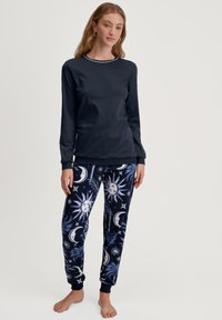 Langschildnavy top met geribbelde halslijn, gecombineerd met joggingbroeken met een patroon van maan- en zonmotieven in blauw en wit op een donkere achtergrond.