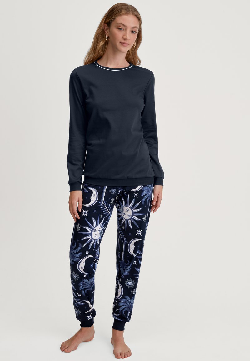 Langschildnavy top met geribbelde halslijn, gecombineerd met joggingbroeken met een patroon van maan- en zonmotieven in blauw en wit op een donkere achtergrond.