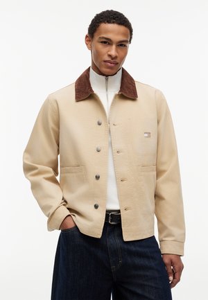 Ung mand iført en beige jakke med knapper og brun krave, hvid lynlås-sweater, mørke jeans, stående med hænderne i lommerne.