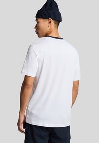 Witte katoenen T-shirt met marineblauwe ribbelkraag, korte mouwen, ontspannen pasvorm, van achteren getoond, in combinatie met donkere shorts en een marineblauwe beanie.