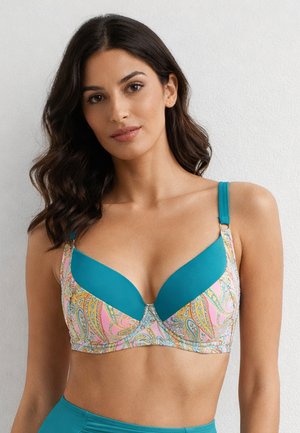 Kobieta o falowanych ciemnych włosach, ubrana w turkusowy top od stanika w wzór paisley, stoi na tle jednolitej jasnej ściany.