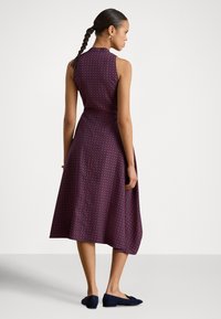 Ermeløs lilla kjole med geometrisk mønster, innsvingt midje og flytende skjørt. Modellen står foran en nøytral bakgrunn iført mørkeblå flate sko.