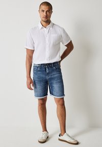 Biała krótkozasięgowa koszula na guziki, niebieskie szorty dżinsowe z podwiniętymi nogawkami i białe sneakersy z beżowymi podeszwami na neutralnym tle.