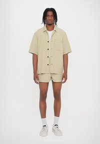 Chemise courte à manches courtes beige avec une poche poitrine, accompagnée de shorts assortis. Portée avec des chaussettes blanches à la cheville et des baskets légères.