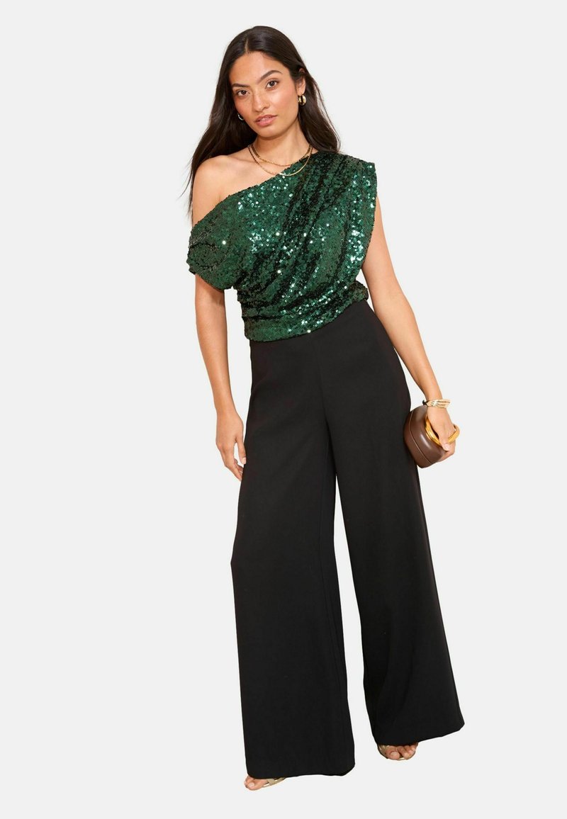 Žalias blizgančiais karoliukais padengtas vieno peties viršus, derinamas su plačiomis juodomis kelnėmis. Modelis laiko rudą rankinę, kuri pasižymi lygiu paviršiumi ir žėrinčiomis detalėmis.