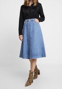 Svart skjorta med fransdetaljer; blå denim A-linjeskirt med framknappar; bruna ormskinnsmönstrade ankelboots med klack.