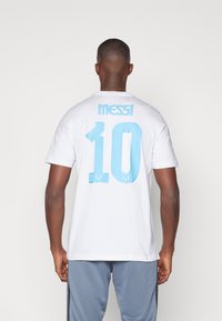 Hvid t-shirt med korte ærmer, hvor der står "MESSI" og nummer "10" trykt i lyseblå på ryggen. Lavet af bomuldsstof, standard pasform, enkelt design.