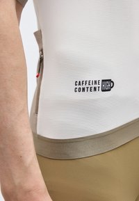 Primo piano del torso che indossa una maglietta bianca a coste con rifiniture beige e il testo "CAFFEINE CONTENT HIGH" accanto a una zip sul lato.