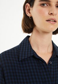 Camisa a cuadros en azul marino con cuello abotonado; presenta una textura suave y un detalle de un solo botón en el cuello. Pendiente en forma de aro dorado visible.
