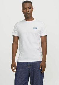 Hvid bomulds t-shirt med rund hals, der har et lille blåt "JACK & JONES" logo på venstre bryst og korte ærmer. Denim jeans bæres nedenunder.