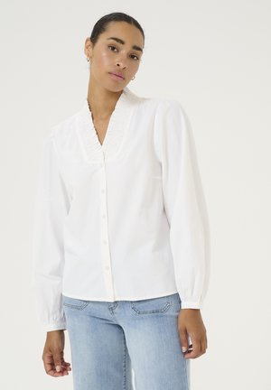 Blusa bianca a maniche lunghe con colletto plissettato, chiusura a bottoni e maniche leggermente a sbuffo. Indossata con jeans di denim azzurri.