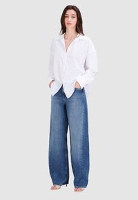 Weißes Baumwollhemd mit Knopfverschluss und Stickereidetails, kombiniert mit weit geschnittenen blauen Denim-Jeans. Die Jeans haben eine ausgebleichte Optik und eine elegante Silhouette.