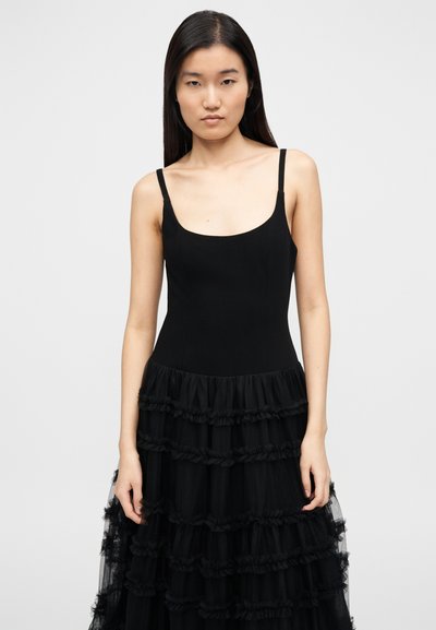 Μαύρο φόρεμα με εφαρμοστή μπούστα και λεπτές τιράντες, καθώς και με πολυεπίπεδη, βολάν φούστα από τούλι. Λείο υλικό στο πάνω μέρος, διάφανα βολάν στο κάτω μέρος.