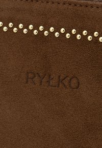 Brūns zamšs ar rindu zelta kniedēm gar augšējo malu un iepresēto logotipu "RYŁKO" centrā. Mīksta materiāla tekstūra un matēta apdare.