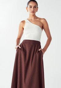 Vrouw in een een-schouder witte ribbed top en een hooggetailleerde bruine rok met haar handen in de zakken, staand tegen een effen witte achtergrond.