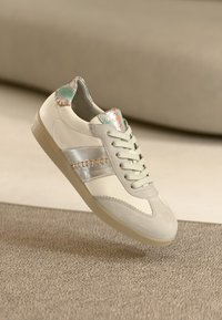 Weißer Ledersneaker mit hellgrauer Wildlederkappe, silbernem Akzentstreifen und mehrfarbig strukturiertem Fersenbereich. Flache Schnürsenkel und Gummisohle.