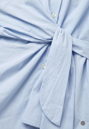 Lichtblauw overhemd met een stoffen riem die in een knoop is gestrikt ter hoogte van de taille, met witte knopen aan de voorkant.