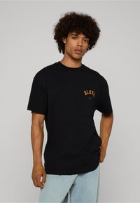 Musta puuvillainen t-paita, jossa on lyhyet hihat ja pyöreä pääntie. Vasemmassa rintapaneelissa oranssilla ja vihreällä brodeerattu "BLKVIS NY".