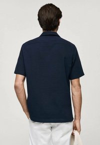 Granatowa koszula krótko rękawowa z kołnierzykiem, wykonana z teksturowanej tkaniny. Posiada schludny design z tyłu oraz wykończenie, zestawiona z jasnymi spodniami.