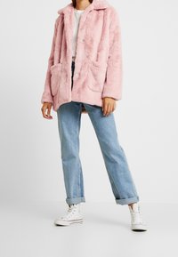 Rosa Kunstpelzmantel mit Knopfverschluss, zwei vorderen Taschen und einem gezackten Kragen, kombiniert mit aufgekrempelten hellblauen Jeans und weißen Turnschuhen.