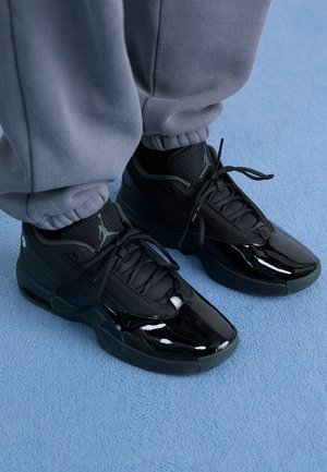Person trägt schwarze glänzende und Mesh-Sneaker mit Schnürsenkeln, steht auf einem hellblauen Teppich, graue Jogginghosen an den Knöcheln hochgekrempelt.