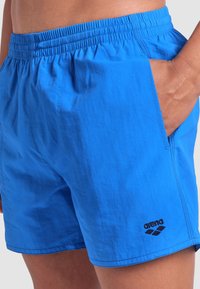 Modré plavky vyrobené z lehkého materiálu, s elastickým pasem, bočními kapsami a černým logem na lemu.