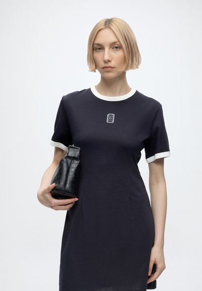 Femme aux cheveux blonds courts portant une robe noire avec une bordure blanche tient un petit sac à main noir texturé sur un fond clair uni.