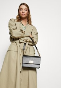 Sort læderhåndtaske med guldlåse, rektangulær form og kædestrop, parret med en beige trenchcoat og mønstret bluse.