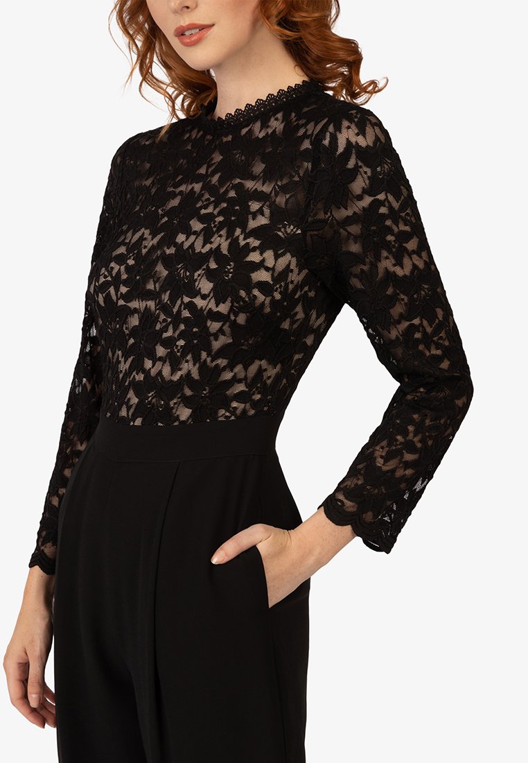 Kobieta w czarnej koronkowej bluzce z długim rękawem w kwiatowe wzory, zestawionej z czarnymi spodniami o wysokim stanie, z ręką w kieszeni.