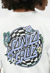 Biely tričko s veľkým grafickým potlačou s textom "Santa Cruz", motýľom, kvetmi a šachovnicovým vzorom v modrej, čiernej a zelenej farbe.