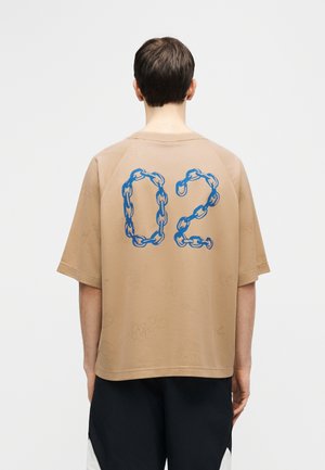 Person wearing a beige oversized t-shirt with blue chain-link style number "02" on the back, paired with black and white pants.