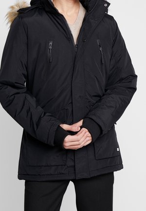 Uomo che indossa una giacca invernale nera con cerniera, tasche con bottoni, polsini a coste e cappuccio bordato di pelliccia, con le mani incrociate davanti.