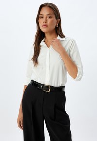 White button-up shirt made of smooth fabric, paired with high-waisted black trousers. An adjustable black patent belt with a gold buckle completes the look.