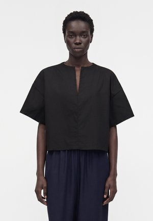 Γυναίκα που φοράει μια άνετη μαύρη κοντομάνικη μπλούζα με μικρή κεντρική σχισμή μπροστά, σε συνδυασμό με σκοτεινό παντελόνι με φαρδιά πόδια, στέκεται μπροστά σε απλό φόντο.