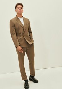 Completo marrone su misura con vita cintata, abbinato a una maglietta bianca e scarpe stringate nere. Tessuto liscio, taglio moderno e vestibilità su misura.