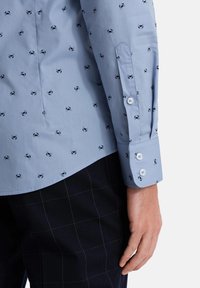 Camisa de color azul claro con un patrón de cangrejos repetido, puños abotonados y una textura suave, combinada con pantalones oscuros a cuadros.