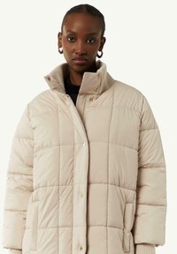 Jasno beżowa kurtka puchowa o pikowanej strukturze, z wysokim kołnierzem, zatrzaskami oraz ribbowymi akcentami na kołnierzu i mankietach.