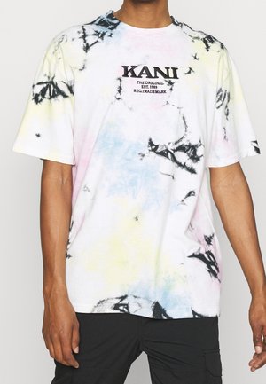 Mand iført løs, kortærmet tie-dye skjorte med teksten "KANI The Original Est. 1989", kombineret med sorte bukser.
