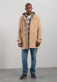 Beige duffelcoat med huva, träknappar som stängning, stora framfickor och ett mjukt, strukturerat tyg, över en rutig skjorta och jeans.