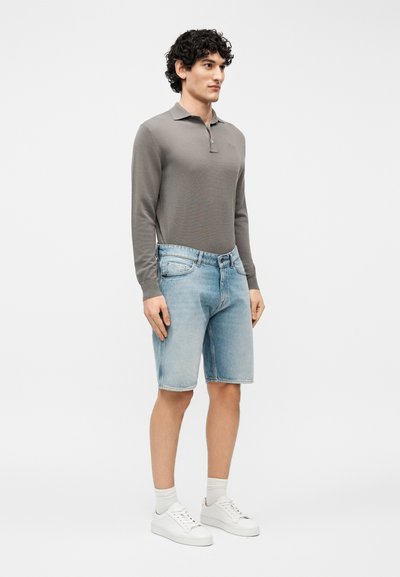 Άνδρας που στέκεται φορώντας γκρι μακρυμάνικο πόλο πουκάμισο, κοντά τζιν σορτς σε ανοιχτό μπλε χρώμα, λευκές κάλτσες και λευκά αθλητικά παπούτσια σε απλό φωτεινό φόντο.