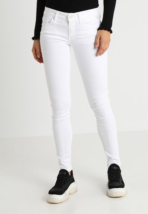 Jeans ajustados blancos con un diseño entallado, estilo de cinco bolsillos y un cierre de botón clásico, combinados con zapatillas negras de suela gruesa.