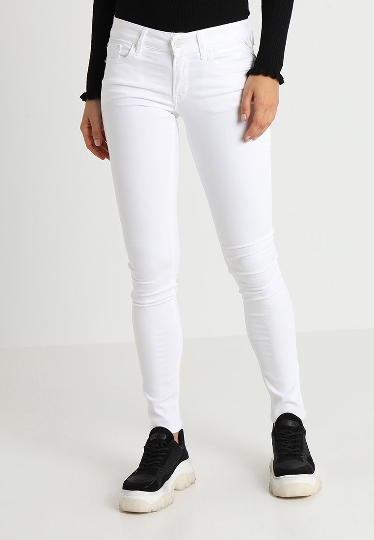 Jeans ajustados blancos con un diseño entallado, estilo de cinco bolsillos y un cierre de botón clásico, combinados con zapatillas negras de suela gruesa.