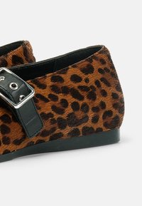 Chaussure plate avec imprimé léopard marron et noir, bride noire avec boucle argentée et semelle noire sur fond blanc.