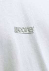 T-shirt en tissu blanc avec un logo en relief texturé de couleur gris clair sur le devant. Présente une texture lisse et un design minimaliste.