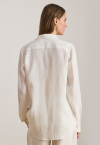 White linen shirt with a classic collar, long sleeves, and a curved hem. Features subtle texture and button detail on the cuffs.