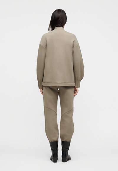 Γυναίκα που στέκεται με την πλάτη, φορώντας ζιβάγκο πουλόβερ ταυπηρής απόχρωσης, ταιριαστό παντελόνι φόρμας με λεπτομέρειες ραφών και μαύρα μποτάκια με τακούνι.