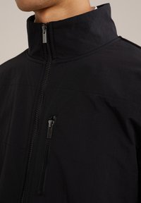 Svart zip-up jacka med hög krage, med en bröstficka på framsidan och textil med struktur. Slät dragkedja och sömdetaljer synliga.