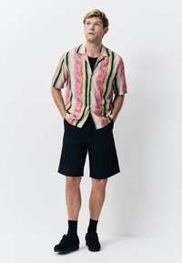 Chemise à manches courtes avec boutons, à rayures verticales roses, vertes et orange, associée à un short noir et des chaussures foncées.