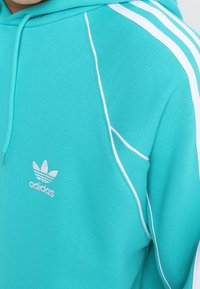 Turkos Adidas hoodie med vit tre-stripes-design och broderad logotyp. Har en huva med dragsko och mjukt, texturerat tyg.