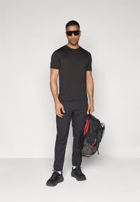 Svart kortermet t-skjorte, svarte bukser og svarte joggesko. Med mørke solbriller, holder en svart og rød ryggsekk med stropper.